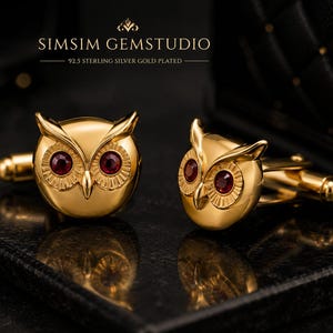 May include: A pair of gold-plated 92.5 sterling silver owl cufflinks featuring deep red circular gemstone eyes. The text SIMSIM GEMSTUDIO and 92.5 STERLING SILVER GOLD PLATED is visible above the owl-shaped accessories on a dark background.