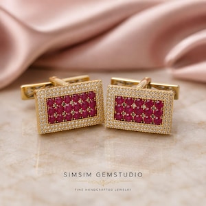 May include: A pair of rectangular gold cufflinks featuring a central cluster of round, deep red rubies surrounded by a border of small, clear white pavé-set stones. The metal frame is polished gold, and the text SIMSIM GEMSTUDIO is visible.