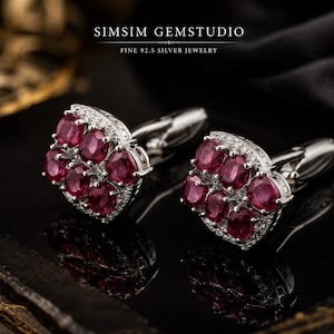 May include: A pair of silver cufflinks featuring a cluster of six oval-cut red rubies surrounded by small white stones. The text SIMSIM GEMSTUDIO FINE 92.5 SILVER JEWELRY is visible at the top. The cufflinks have a polished silver finish.