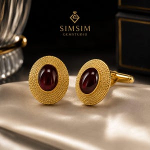 Puede incluir: Un par de gemelos dorados con una piedra preciosa cabujón de color rojo intenso de forma ovalada en el centro. El borde circundante tiene un patrón texturizado similar a una cuerda. El texto SIMSIM GEMSTUDIO es visible arriba.