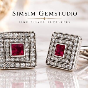 Op de afbeelding: Een paar vierkante zilveren manchetknopen met een centrale prinsesgeslepen rode edelsteen, omringd door twee concentrische randen van kleine heldere steentjes. De tekst SIMSIM GEMSTUDIO FINE SILVER JEWELLERY staat boven de manchetknopen.