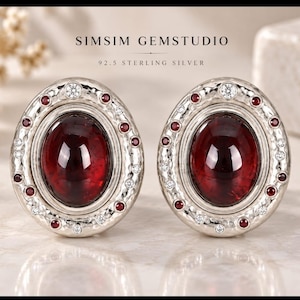 Peut inclure: Une paire de boucles d'oreilles ovales en argent sterling 92.5 de SIMSIM GEMSTUDIO, chacune dotée d'une grosse pierre grenat cabochon centrale entourée d'un bord en argent martelé orné de petits grenats rouges et de pierres blanches.