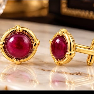 May include: A pair of gold-toned cufflinks featuring a circular design with a polished, deep red cabochon stone at the center. The metal frame has a twisted rope-like texture around the perimeter, and the toggle mechanism is visible on the side.