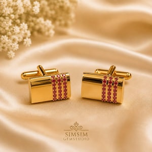 May include: A pair of gold-toned rectangular cufflinks featuring three vertical rows of small, round, vibrant red gemstones set into the polished metal surface. The cufflinks have a classic T-bar fastening mechanism on the back for secure attachment.