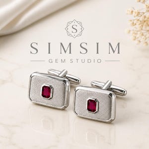 Könnte beinhalten: Ein Paar silberne Manschettenknöpfe mit rechteckigem Design, jeweils mit einem zentralen, tiefroten Edelstein. Die Manschettenknöpfe werden auf einer hellen Oberfläche präsentiert, mit dem Text "SIMSIM GEM STUDIO" darüber.