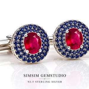Puede incluir: Un par de gemelos de plata de ley con una piedra roja central de corte ovalado rodeada por un doble halo de pequeñas piedras azules. El texto SIMSIM GEMSTUDIO 92.5 STERLING SILVER está impreso debajo de los gemelos sobre fondo blanco.