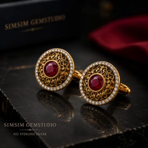 May include: A pair of round cufflinks featuring a central faceted red stone surrounded by intricate gold-toned filigree metalwork and a border of small clear stones. The text SIMSIM GEMSTUDIO and 925 STERLING SILVER is visible in the image.