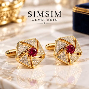 Könnte beinhalten: Ein Paar goldfarbene Manschettenknöpfe mit geometrischem, fünfeckigem Design, besetzt mit kleinen weißen Steinen und einem zentralen runden roten Edelstein. Der Text SIMSIM GEMSTUDIO ist darüber zu sehen. Die Metalloberfläche ist poliert und glänzend.