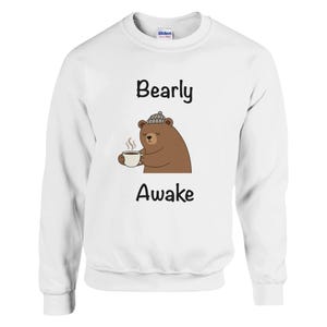Peut inclure: Sweat-shirt blanc avec l'inscription "Bearly Awake". Un ours de dessin animé portant un chapeau gris tient une tasse de café. L'ours est brun avec un ventre brun clair.