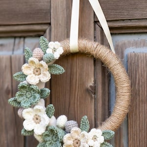 Puede incluir: Una corona hecha a mano con una base envuelta en yute, adornada con flores y hojas de ganchillo en crema, beige y verde. Se utiliza una cinta color crema para colgarla. La corona se exhibe contra una puerta de madera.