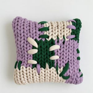 Puede incluir: Un cojín decorativo con un diseño de punto grueso. El cojín presenta un patrón de patchwork con secciones de hilo lavanda, crema y verde. Las puntadas de color crema y los detalles verdes en forma de hoja añaden detalles.