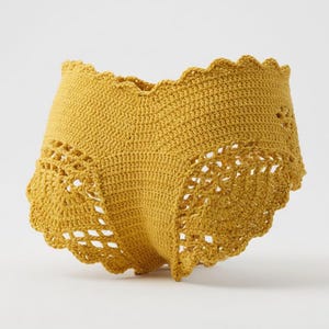 May include: Mustard yellow crochet shorts with a scalloped edge. The shorts feature a textured, openwork design on the sides. The handmade garment is displayed against a white background.