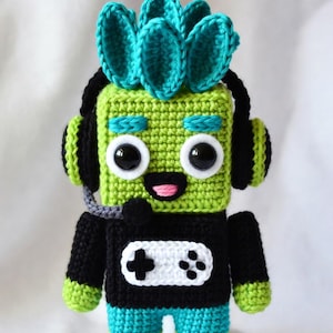 Puede incluir: Muñeco de ganchillo con temática de jugador. El muñeco tiene una cabeza cuadrada verde con ojos grandes, un mohawk turquesa y auriculares negros. Lleva una camiseta negra con un diseño de mando de juego blanco y pantalones cortos turquesa. El muñeco mide unos 15 cm de alto.