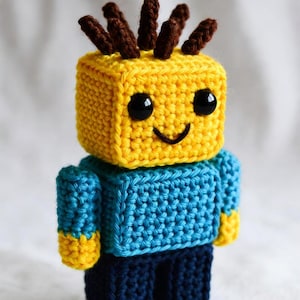 Puede incluir: Muñeca de ganchillo hecha a mano con una cabeza cuadrada amarilla y una cara sonriente. La muñeca tiene un cuerpo azul, pantalones azul marino y cabello castaño. Los brazos y los pies de la muñeca son amarillos.