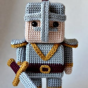 Puede incluir: Figura de caballero tejida a mano en tonos grises, rosas y dorados. El caballero lleva un casco, hombreras y un cinturón. Sostiene una espada con empuñadura dorada y hoja burdeos. Ojos negros de botón.