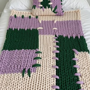 以下が含まれることがあります： パッチワークデザインのチャンキーニットブランケットと枕のセット。ブランケットはクリーム、グリーン、ラベンダーのセクションがあり、装飾的なステッチが施されています。枕はブランケットのデザインを反映しています。白いベッドにセットが飾られています。