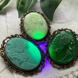 Puede incluir: Tres broches de camafeo ovalados en tonos verdes y blancos, cada uno con un diseño de perfil detallado. Los broches tienen marcos ornamentados de color bronce. El broche central brilla con una luz verde brillante, mientras que los otros tienen un brillo sutil.