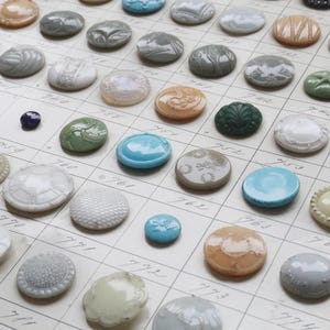 Puede incluir: Una colección de botones vintage en varios colores y diseños, incluyendo formas redondas, ovaladas y florales. Los botones están dispuestos en una cuadrícula, mostrando sus diversas texturas y tamaños. Los colores van del azul y verde al blanco y naranja.