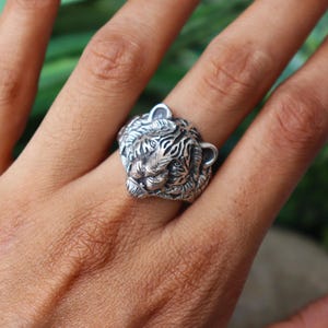 Könnte beinhalten: Ein detaillierter Silberring in Form eines Tigerkopfes. Der Ring weist aufwendige Schnitzereien auf, die die Gesichtszüge des Tigers hervorheben, darunter seine Streifen, Nase und Ohren. Der Ring wird an einem Finger getragen und zeigt sein Design.