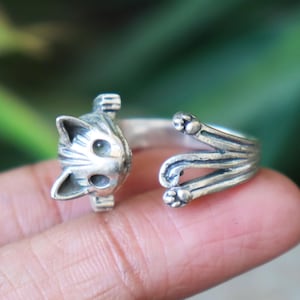 Könnte beinhalten: Ein silberner Katzenring. Der Ring hat einen Katzenkopf auf der einen Seite und Pfoten auf der anderen Seite, wodurch ein offenes Design entsteht. Das Gesicht der Katze hat detaillierte Merkmale, einschließlich Augen und Ohren. Der Ring wird an einem Finger getragen.