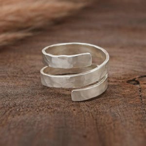 Könnte beinhalten: Ein silberfarbener Ring mit gehämmertem Muster in Spiralform. Der Ring umschließt den Finger mit einer strukturierten Oberfläche und erzeugt einen modernen Look. Der Ring wird auf einer Holzoberfläche präsentiert.