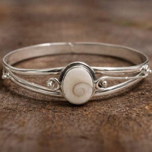 Pode incluir: Uma pulseira de prata com uma peça central ovalada esbranquiçada. A peça central apresenta um desenho em espiral. A pulseira tem redemoinhos decorativos e está sobre um fundo de madeira. Uma joia.
