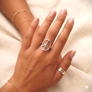 Könnte beinhalten: Ein silberner Ring mit einem Friedenssymbol. Der Ring wird an einer Hand getragen, mit zwei weiteren Ringen an einem anderen Finger. Auch ein goldenes Armband ist zu sehen. Ein Schmuckstück, das Frieden symbolisiert.