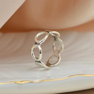 Könnte beinhalten: Ein silberner Ring mit offenem Oval-Design. Der Ring besteht aus glänzendem Metall und weist eine Reihe von miteinander verbundenen Ovalen auf. Der Ring wird auf einer weißen Oberfläche mit goldenem Rand vor einem weichen, neutralen Hintergrund präsentiert.