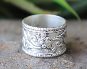 May include: A silver-colored ring with an intricate floral design. The ring features a band with embossed flowers and swirling patterns. The ring is approximately 1 inch wide and has a textured surface.