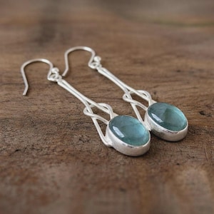 Könnte beinhalten: Ein Paar silberfarbene Ohrhänger mit ovalen, hellblauen Edelsteinen. Die Ohrringe haben ein einzigartiges Design mit einer Schlaufendetail über dem Edelstein. Die Edelsteine sind in einer silbernen Lünette gefasst.