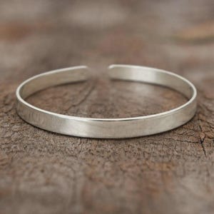 Könnte beinhalten: Ein silbernes Armreif-Armband mit schlichtem, minimalistischem Design. Das offene Armband besteht aus glattem, glänzendem Metall und liegt auf einer strukturierten Holzoberfläche. Das Armband ist etwa 0,6 cm breit.