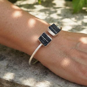 Könnte beinhalten: Ein silberfarbenes, offenes Armband mit zwei quadratischen Fassungen an den Enden. Jede Fassung enthält einen rauen, vertikal gestreiften schwarzen Stein. Das Metallband hat eine matte Oberfläche und ein schlichtes, offenes Design.