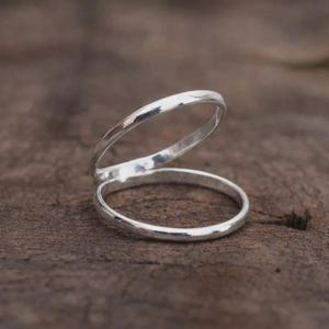 Könnte beinhalten: Ein silberner Doppelring mit minimalistischem Design. Die beiden Bänder sind verbunden und schaffen einen einzigartigen und modernen Look. Der Ring ist vor einem rustikalen Holzgrund platziert, der sein poliertes Finish hervorhebt.
