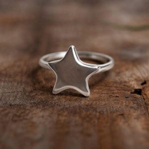 Könnte beinhalten: Ein silberner Ring mit einem sternförmigen Design. Der Stern ist das Hauptmerkmal, das auf einem schlanken, polierten Band sitzt. Der Ring wird auf einer strukturierten Holzoberfläche gezeigt.