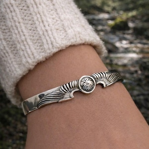 Könnte beinhalten: Ein silbernes Armreif-Armband mit detailliertem Sonnenmuster. Das Armband hat ein zentrales kreisförmiges Emblem mit einem Sonnenmotiv, flankiert von stilisierten flügelartigen Mustern. Der Schmuck wird am Handgelenk getragen, vor dem Hintergrund eines cremefarbenen Pullovers.