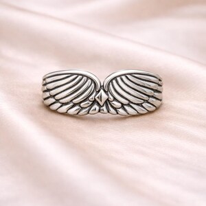 Peut inclure: Une bague argent&eacute;e orn&eacute;e d'ailes sym&eacute;triques. Les ailes de la bague sont d&eacute;taill&eacute;es avec de multiples lignes, et un accent en forme de losange est plac&eacute; au centre. La bague est pr&eacute;sent&eacute;e sur un tissu rose clair et doux.