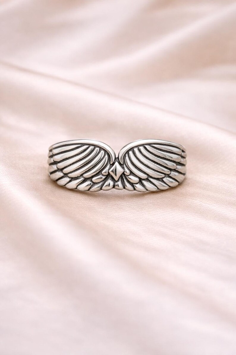 Peut inclure: Une bague argent&eacute;e orn&eacute;e d'ailes sym&eacute;triques. Les ailes de la bague sont d&eacute;taill&eacute;es avec de multiples lignes, et un accent en forme de losange est plac&eacute; au centre. La bague est pr&eacute;sent&eacute;e sur un tissu rose clair et doux.