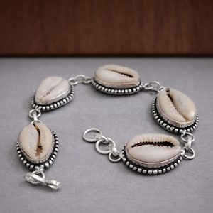 Könnte beinhalten: Ein silbernes Armband mit sechs natürlichen Kaurischnecken. Jede Muschel ist in einer silbernen Lünette mit einem Perlenrand gefasst und durch silberne Glieder verbunden. Das Armband hat einen Verschluss.