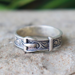 Könnte beinhalten: Ein silberfarbener Ring mit Schnallen-Design. Der Ring hat ein eingraviertes Muster um das Band und eine detaillierte Schnalle. Der Ring ist vor einem neutralen, strukturierten Hintergrund platziert.
