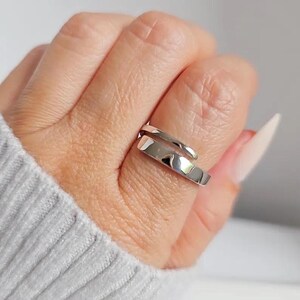 Könnte beinhalten: Ein silberfarbener Ring mit modernem, minimalistischem Design. Der Ring umschließt den Finger mit einem schlanken, überlappenden Band. Das polierte Metall reflektiert das Licht und erzeugt ein helles, auffälliges Aussehen. Der Ring ist ein Schmuckstück.