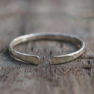 Könnte beinhalten: Ein silberfarbenes Armreif-Armband mit gehämmertem Muster. Das offene Design hat flache, abgerundete Enden. Das Armband wird auf einer verwitterten Holzoberfläche gezeigt, was seinen handwerklichen Charakter unterstreicht. Durchmesser ca. 6,5 cm.