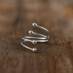 Könnte beinhalten: Ein silberfarbener Ring mit einem Spiraldesign. Der Ring hat vier kleine, runde, kugelförmige Akzente, die gleichmäßig über die Spirale verteilt sind. Der Ring ist auf einer warmen, braunen Holzoberfläche platziert.