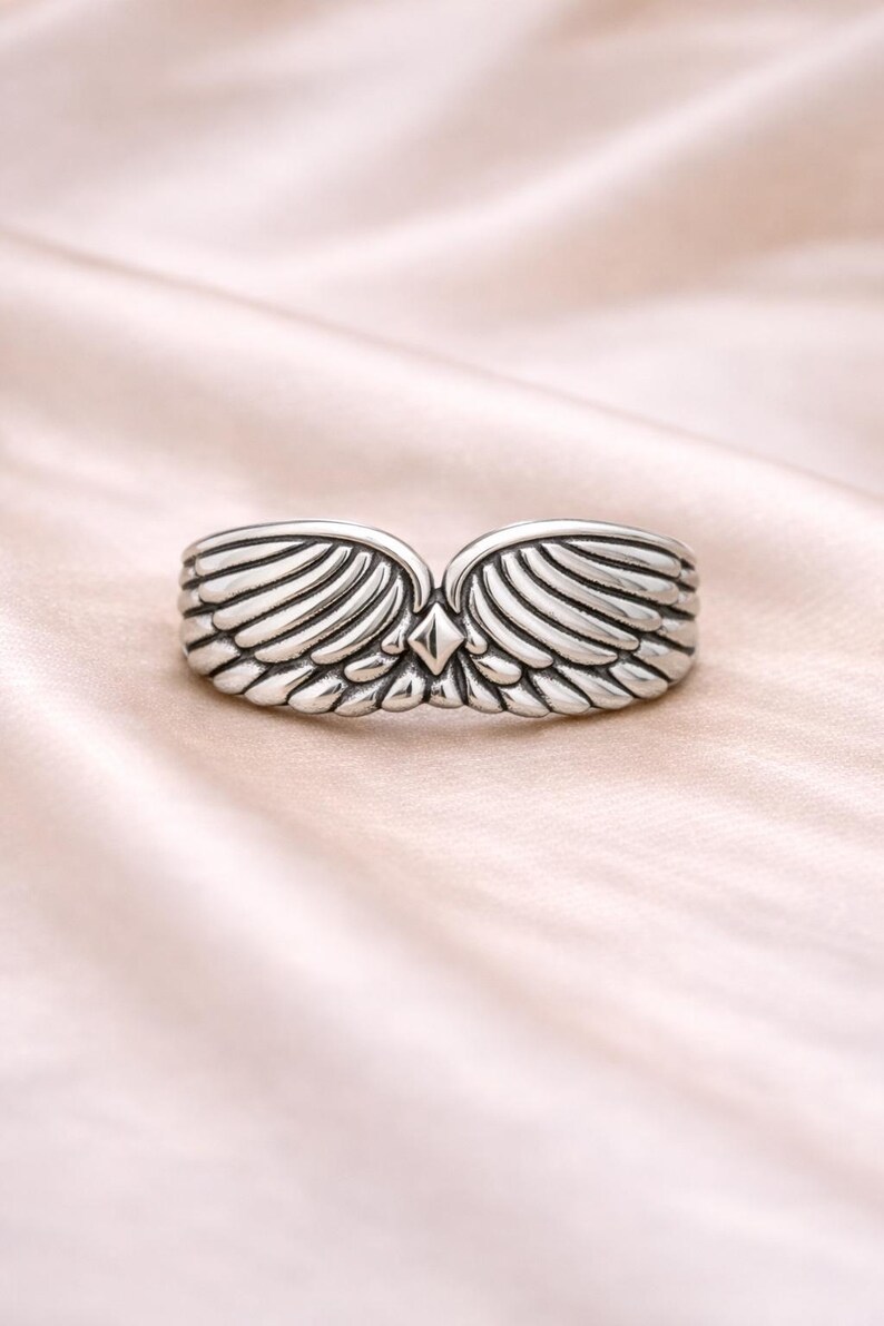 Peut inclure: Une bague argent&eacute;e avec un motif d'ailes. La bague pr&eacute;sente deux ailes d&eacute;taill&eacute;es qui se rejoignent au centre, avec un accent en forme de losange. La bague est pos&eacute;e sur un fond rose doux.