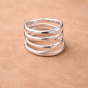 Könnte beinhalten: Ein silberfarbener Ring mit einem Spiraldesign. Der Ring besteht aus fünf verbundenen Bändern, die sich umeinander wickeln. Der Ring wird vor einem neutralen Hintergrund gezeigt.