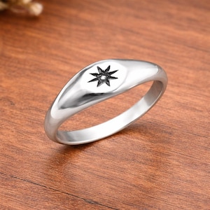 Könnte beinhalten: Ein silberner Ring mit einem schwarzen Sternmotiv. Der Ring hat ein glattes, poliertes Band und eine leicht ovale Oberfläche. Der Stern ist in der Mitte der Ringoberfläche platziert. Der Ring liegt auf einer Holzoberfläche.