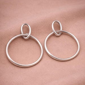 Könnte beinhalten: Ein Paar silberne Creolen. Jeder Ohrring hat einen großen, kreisförmigen Reifen, der oben mit einer kleineren, ovalen Schlaufe verbunden ist. Die Ohrringe sind vor einem weichen, rosa Hintergrund abgebildet.
