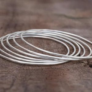 Könnte beinhalten: Eine Ansammlung dünner, silberfarbener Armreifen auf einer rustikalen, braunen Holzoberfläche. Die Armreifen sind kreisförmig und scheinen aus Metall zu sein. Der Hintergrund ist eine verschwommene, strukturierte Holzmaserung.