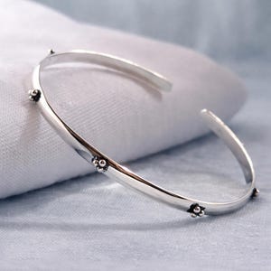 Könnte beinhalten: Ein silberfarbenes Armreif-Armband mit minimalistischem Design. Das Armband hat kleine, dekorative Blumenakzente, die gleichmäßig über das Band verteilt sind. Das offene Design ermöglicht ein einfaches Tragen. Das Armband wird auf einer weißen Oberfläche präsentiert.