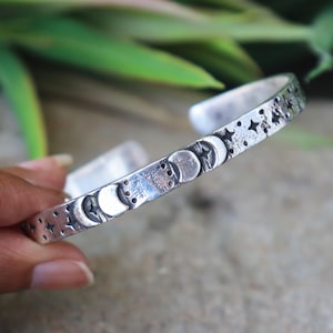 Könnte beinhalten: Ein silberfarbenes Metall-Armband in offener Form mit oxidiertem Finish, das mit einem wiederkehrenden Muster aus Mondsicheln, kreisförmigen Mondphasen und vierzackigen Sternen auf der Außenseite des Bandes graviert ist.