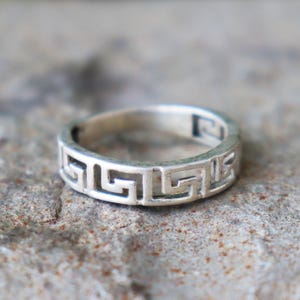 May include: A silver ring with a Greek key design. The ring has a circular shape and a repeating geometric pattern around the band. The ring's metallic sheen and intricate details are highlighted against a neutral, textured background.